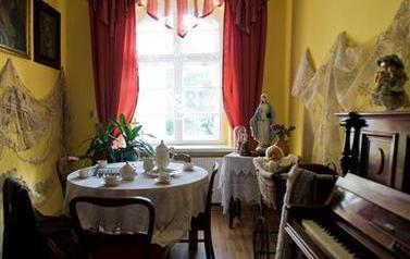 Fotografia przedstawia wnętrze pokoju gościnnego znajdującego się w Izbę Regionalną w Przyszowicach. St&oacute;ł przykryty białym obrusem a nim zastawa porcelanowa. Obok stoi pianino. Na ścianach wiszą obrazy.