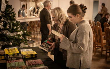 Na pierwszym planie kobiety stoją przed stoiskiem z książkami i czytają ksiazki.